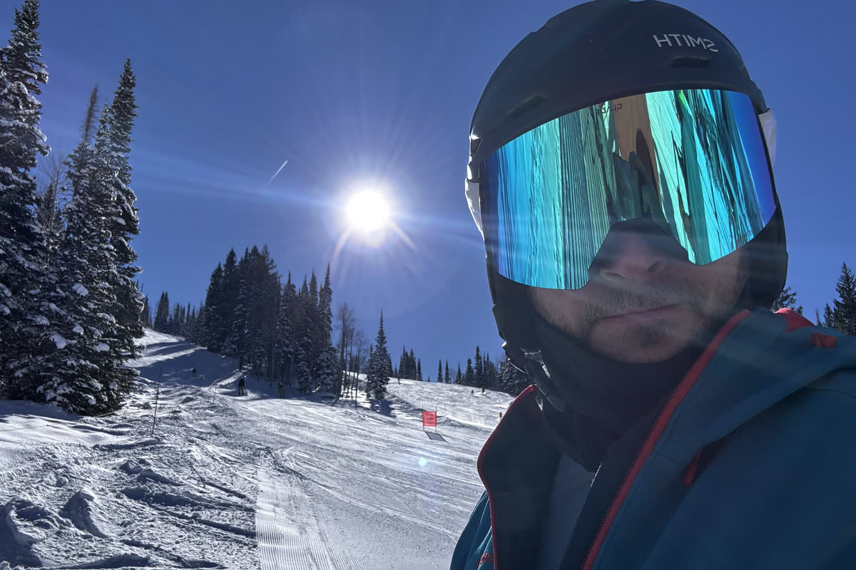 Selfie of Goody in a helmet and mirrored goggles beside a freshly groomed run, snowy pines and the sun in a deep blue sky.