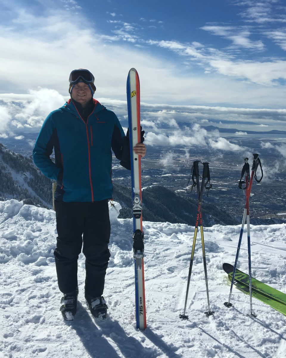 Goody on a snowy summit at Snowbasin holding a vintage red, white and blue K2 715 ski, poles planted in the snow and the valley below the clouds.
